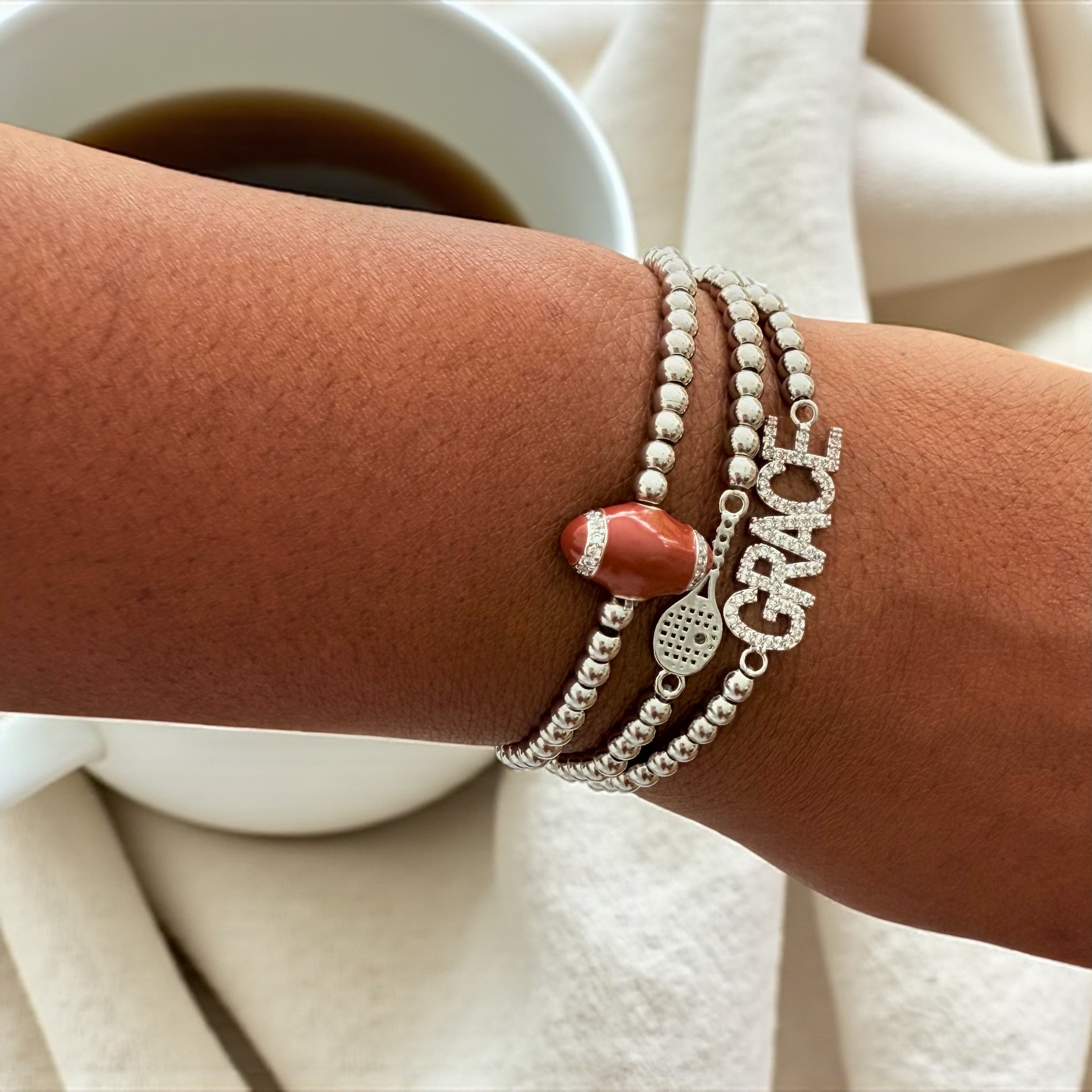Stack of beaded bracelets with a football charm, a tennis racket charm, and a "GRACE" charm.